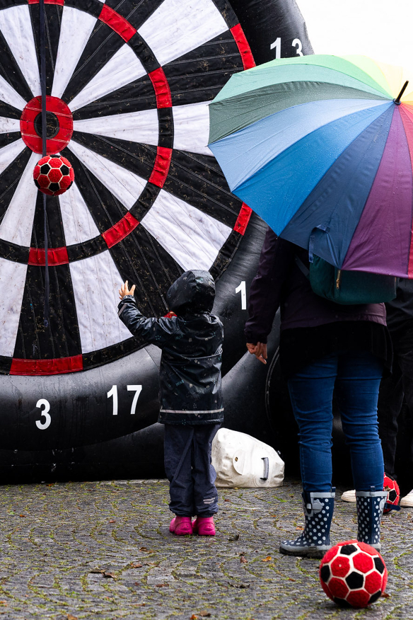 Ein Kind wirft einen Ball auf eine riesige Dartscheibe, es ist ein bunter Regenschirm zu sehen - Copyright: Niko Spettmann